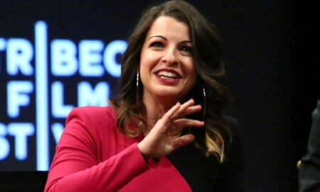 Anita Sarkeesian, in a black and pink dress, speaks at an event.