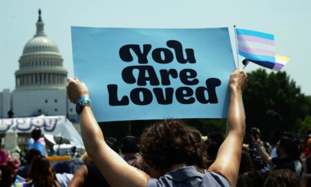 A person holds up two trans flags and a sign reading