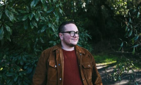 Trans, non-binary rabbi Elliot Kukla wears a red shirt and brown coat as he stands outside near some trees