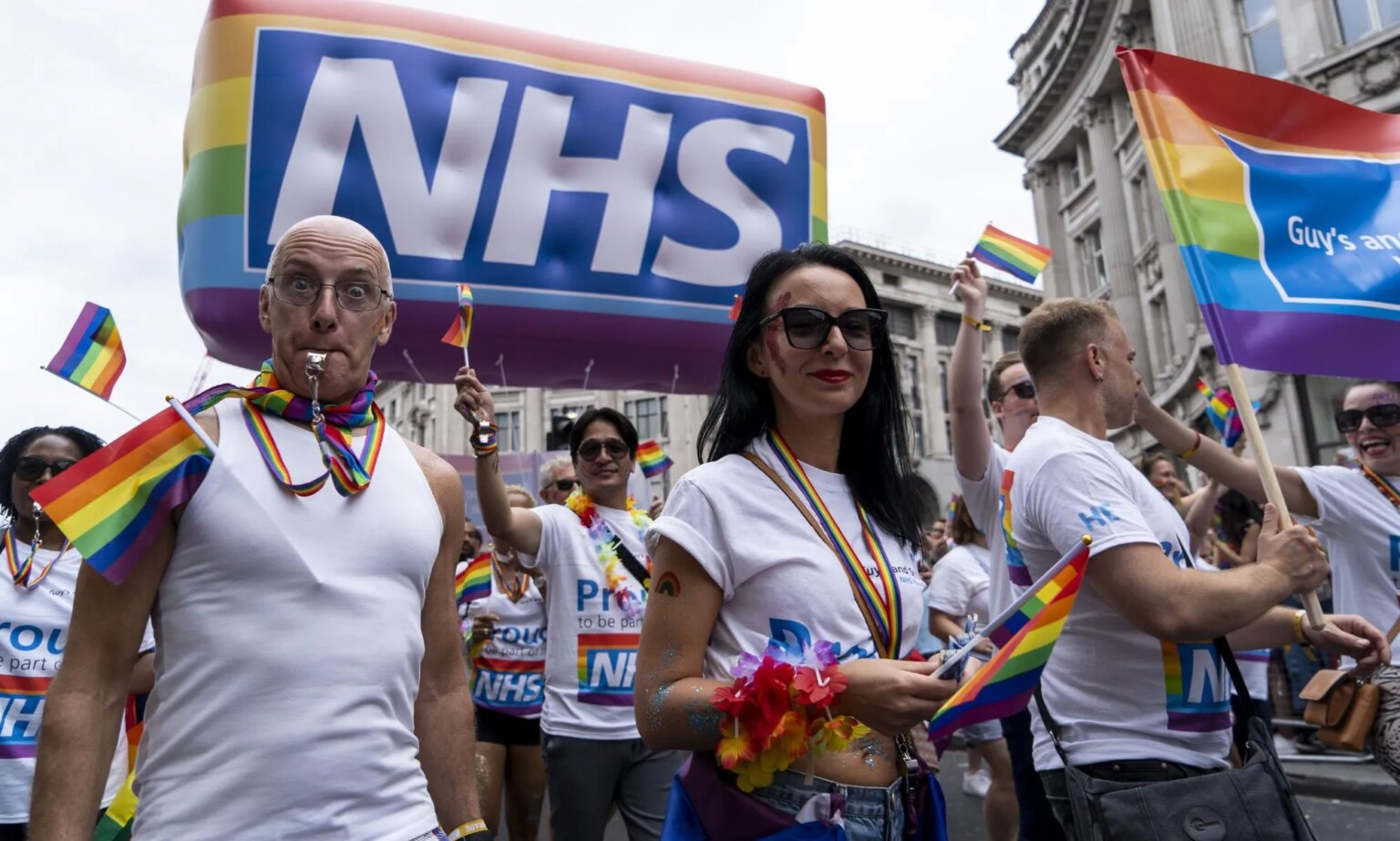 Les personnes trans auraient attendu jusqu’à 35 ans pour un premier rendez-vous sur le NHS Protestors wearing rainbow accessories stand below an NHS Pride balloon.