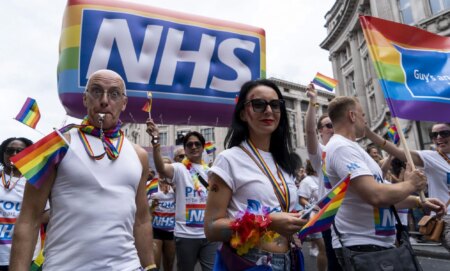 Protestors wearing rainbow accessories stand below an NHS Pride balloon.