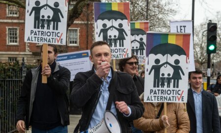 A man holds a megaphone while people behind him hold up anti-LGBTQ+ Patriotic Alternative signs.