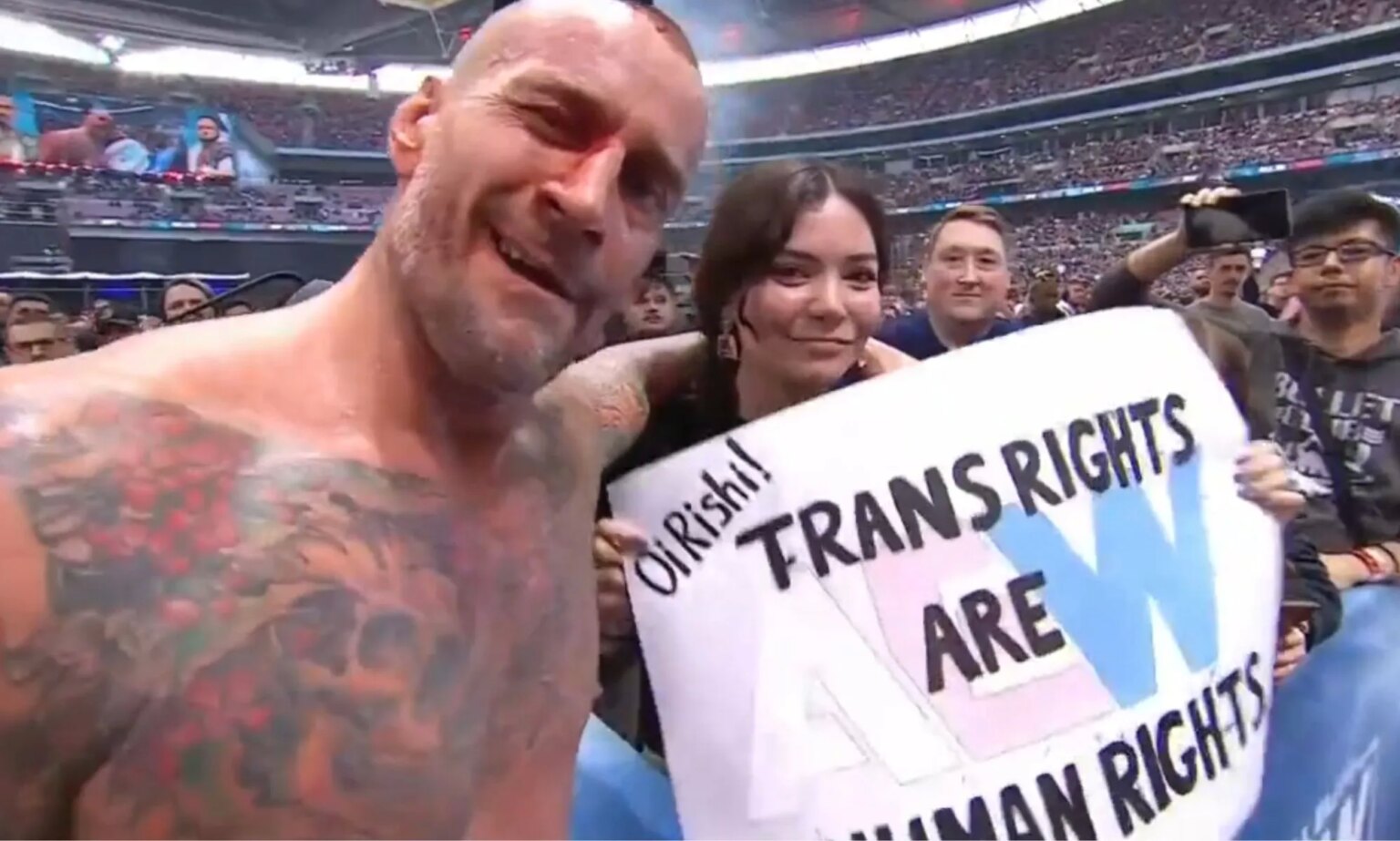 CM Punk pose avec la pancarte d’un fan pour les droits des trans appelant Rishi Sunak CM Punk with a fan holding a sign that reads