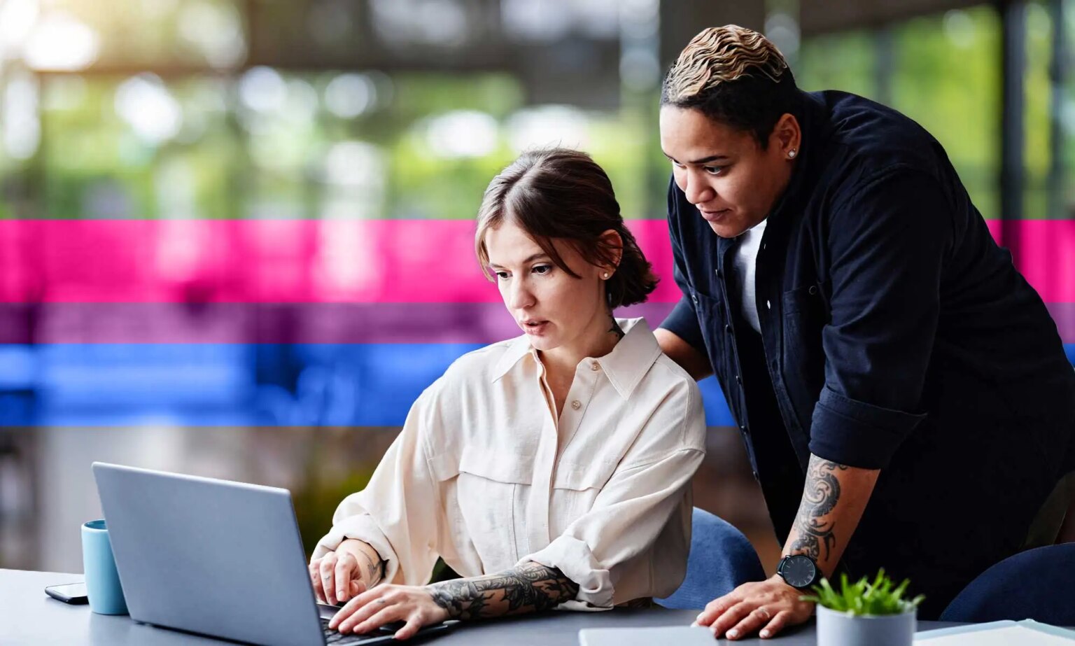 L’effacement des personnes bisexuelles est également un problème sur le lieu de travail – voici comment le combattre This is an image of two female presenting people at work. One person is sitting at a desk looking at a computer and they are looking at a computer. The other person is standing over them.