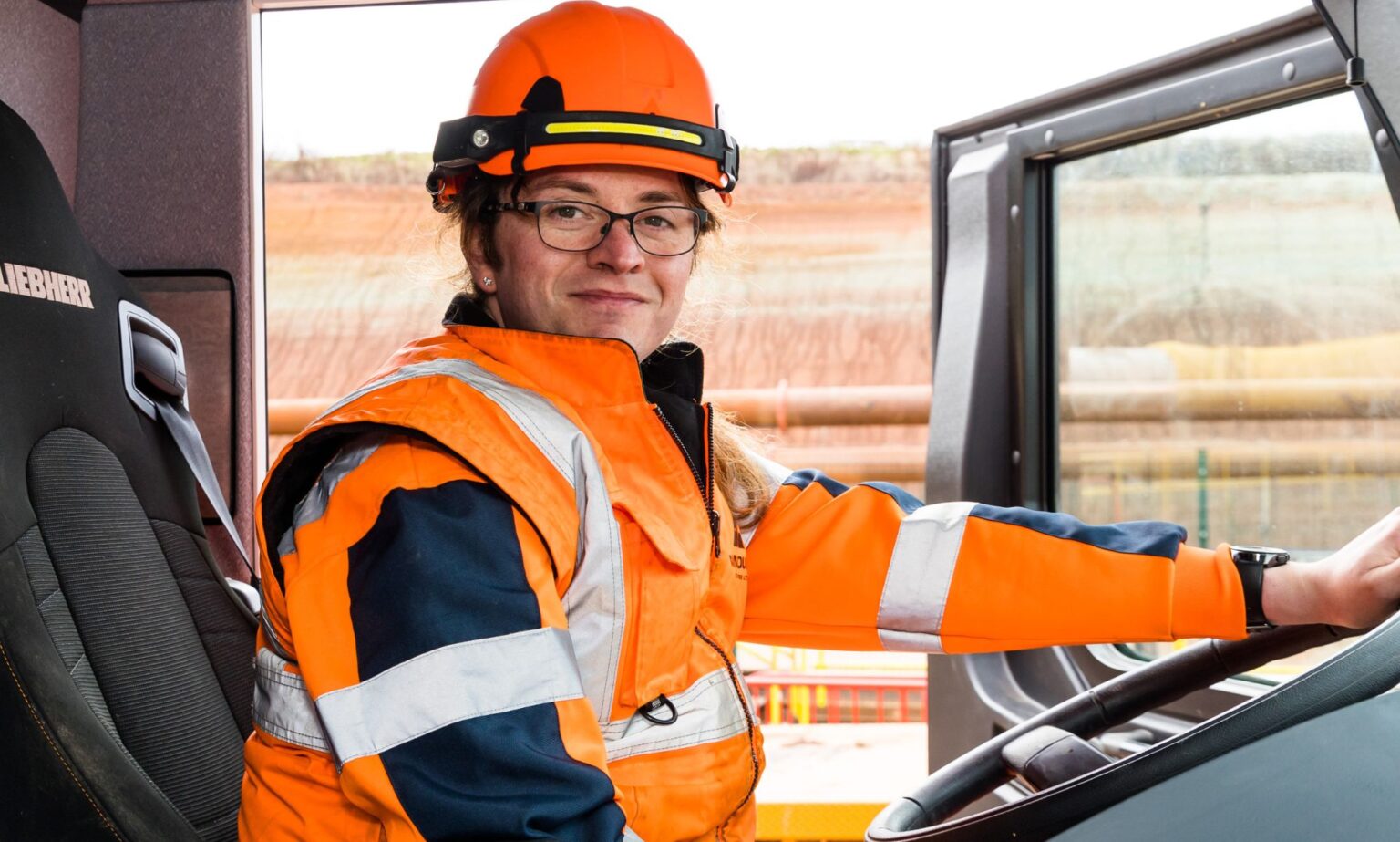 La grutier Trans HS2 parle de son retour au travail « validant » après la transition This is an image of a trans woman and HS2 worker, Kat, sitting inside a truck. She is wearing an orange hi-vis jacket and helmet. The door of the cab is open and she has one hand on the steering wheel.
