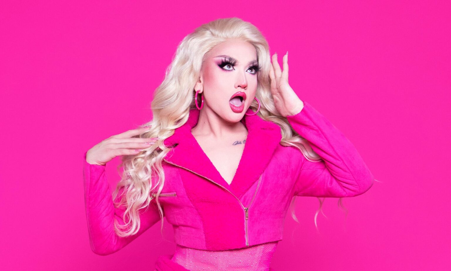 Lagoona Bloo veut devenir la première drag pop star à figurer en tête des charts : « Je me vois aux Grammys » Drag star Lagoona Bloo in a pink jacket looking surprised holding her hand to her forehead. She is stood against a hot pink background.