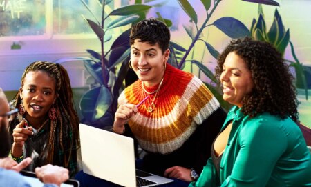 This is an image of a diverse goup of colleagues. On the left there is a Black woman with long braids and a black jumper. In the middle is a white woman with short hair sitting over a laptop. The woman on the right has dark hair and is wearing green.