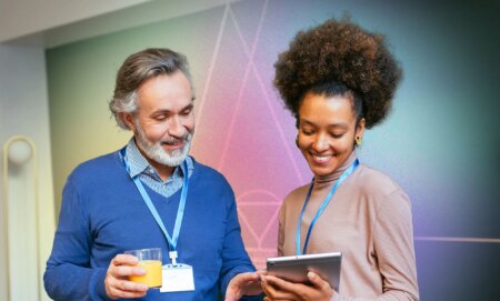 This is an image of two people looking at an iPad, the man on the left is wearing blue. The woman on the right is Black and is wearing a pinkish jumper.