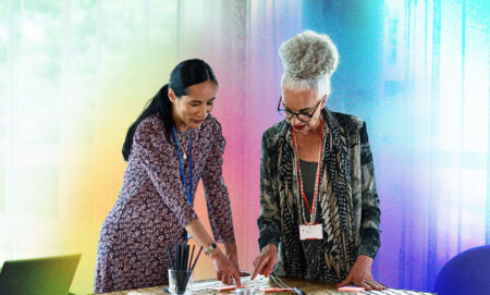 This is an image of two women. The woman on the left is wearing a dress and pointing down at a table. The other woman is wearing a green floral print blazer