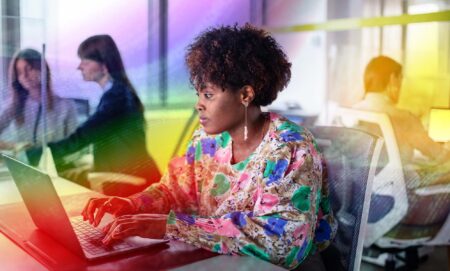 This is an image of a Black woman working at a computer. She is wearing a floral top. There is a creative overlay with the Pride colours over the top