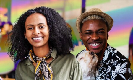 This is an image of two young Black people. The person on the left is a woman and she has long black hair. She is wearing a green top with a multicoloured scarf. The person on the right is male presenting, with a hat on and a floral shirt. He is holding a dog.