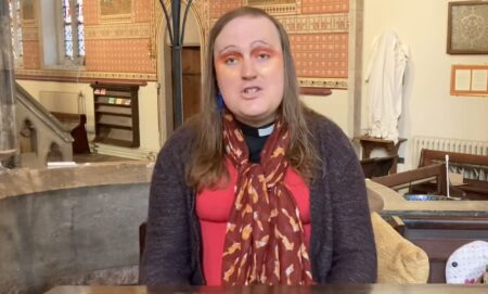 Bingo Allison, a non-binary, genderqueer Church of England priest, wears a red top, sweater and scarf while they speak about religion and Trans Day of Visibility