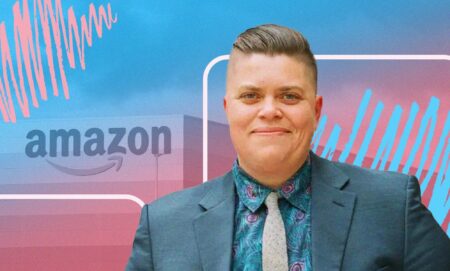 This is a picture of a trans man. He is wearing a bluish grey suit. In the background there is a logo of Amazon with a creative overlay using the colours of the trans flag, pink, white and blue