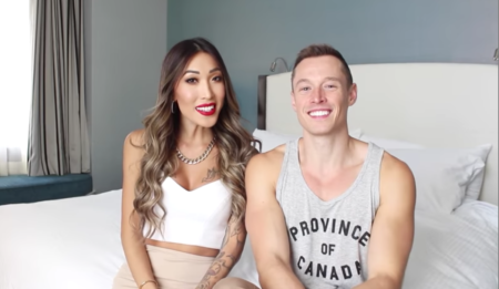 Two friends - a gay youtuber in a tank top and a trans woman in a white vest top - sit together on a bed.