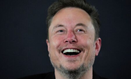 Elon Musk laughing infront of a grey background.