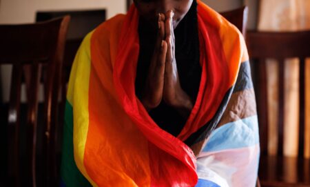 A person in Uganda wears an LGBTQ+ Progressive Pride flag draped around their shoulders as they hold their hands in front of them in prayer