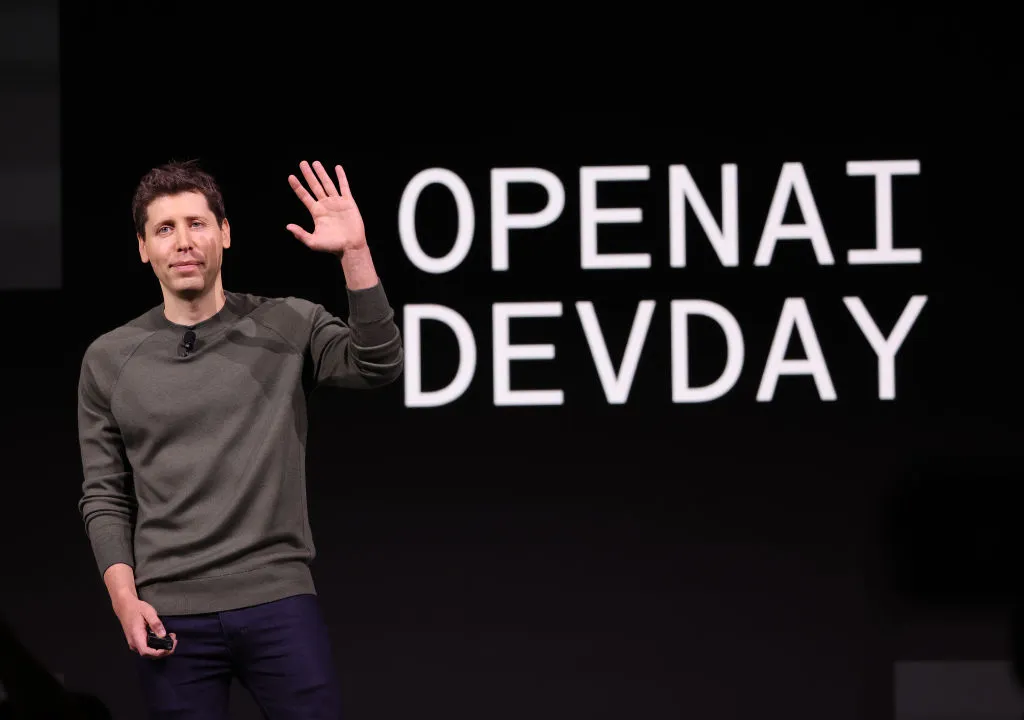 Ceci est une photo de Sam Altman, le PDG d'OpenAI.  Il est debout sur une scène et fait signe de la main droite. 
 