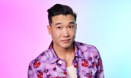 Joel Kim Booster in a jacket with purple faces and red lips. The background is hazy pink and blue.