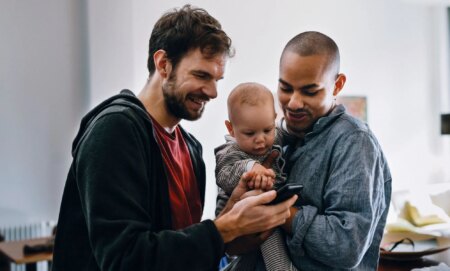 Two men holding a baby watch a video on their mobile phone.