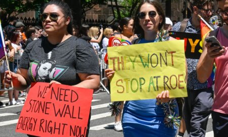 New York City Pride parade with one person holding a sign that says 'We need Stonewall energy right now' and another person holding a sign that says 'They won't stop at Roe'