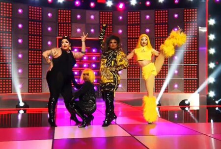 Image shows four Drag Race queens on stage doing the girl group challenge, wearing flamboyant costumes. One is high kicking