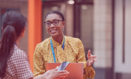 This is an image of a Black woman speaking with a colleague. She is wearing glasses and smiling.