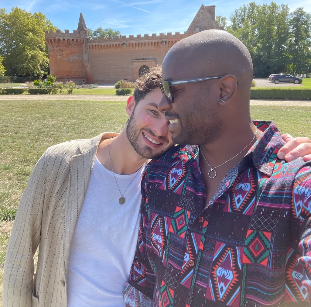 Il s'agit d'une image de deux hommes s'embrassant.  L'homme de gauche est blanc et porte un cardigan beige et un t-shirt blanc.  L'homme de droite est noir et porte une chemise à imprimé aztèque et des lunettes de soleil foncées. 