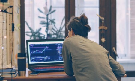 This is an image of a person working at a computer. They are facing away from the camera. There looks to be financial market information on their computer screen.