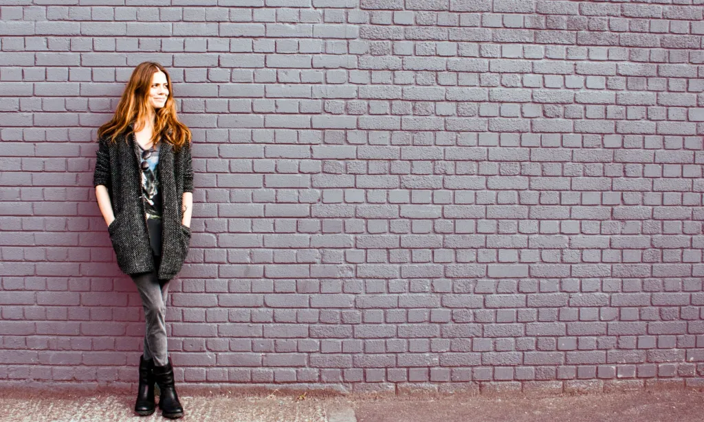Il s'agit d'une photo d'une femme appuyée contre un mur.  Elle a les cheveux roux et porte du noir