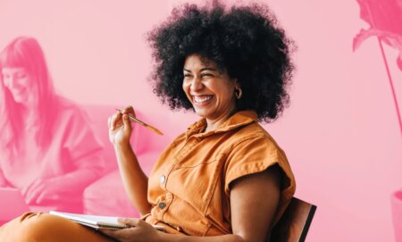 This is an image of a Black woman wearing an orange jumpsuit. She is in a work meeting and is smiling. She is above a pink backgroud.