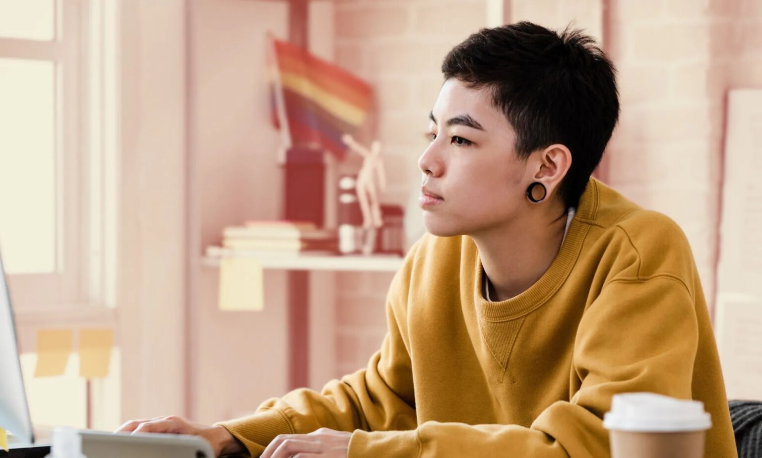 Le personnel LGBTQ+ de la génération Z déclare qu'il ne se sent pas inclus au travail – les employeurs pourraient en perdre des millions This is an image of a person working at a computer. They present genderqueer. They have large gauge earrings and are wearing a yellow jumper.