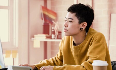 This is an image of a person working at a computer. They present genderqueer. They have large gauge earrings and are wearing a yellow jumper.