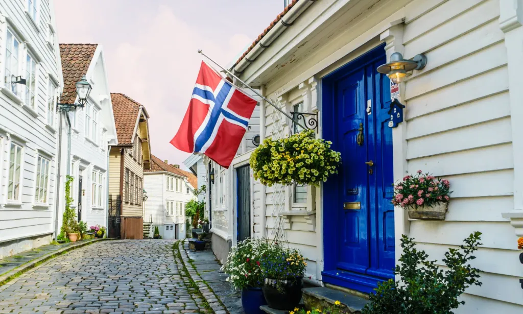 Il s'agit d'une image du drapeau norvégien dans une rue.  Le drapeau est rouge avec une croix bleue aux contours blancs et il est accroché devant une porte bleue dans une petite rue du village.