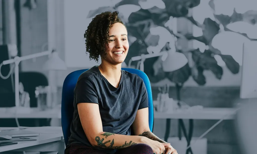 Il s’agit d’une image d’une femme se présentant assise sur une chaise.  Ils portent un t-shirt sombre et portent divers tatouages ​​visibles.  Elle sourit.  Elle est en couleur tandis que le reste de l'image est en noir et blanc. 