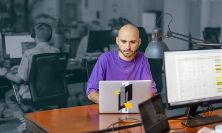 This is an image of a man working at a laptop computer. He is bald and is wearing a purple t shirt. He is in full colour and the rest of the image is in black and white.