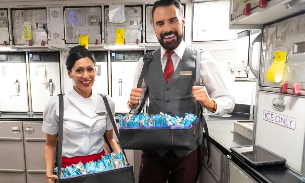 Il s'agit d'une image de la célébrité Rylan Clark debout à côté d'une femme dans la cuisine d'un avion.  Ils portent l'uniforme du personnel de cabine de Virgin Atlantic et disposent de plateaux de collations.