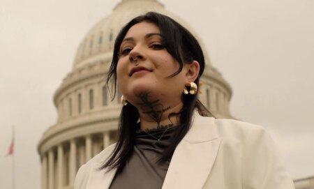 Non-binary activist Leah Juliett looks down at the camera while wearing a dark shirt and white blazer while standing in front of a US historic building