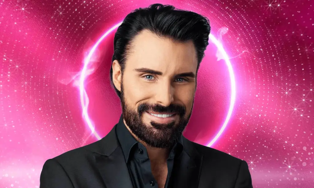 C'est une image de Rylan Clark. Il a les cheveux foncés, une barbe courte. Il porte un costume noir et sourit.