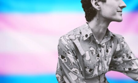 a graphic composed of the blue, pink and white stripes of the trans flag and a masculine person sitting as they look off camera