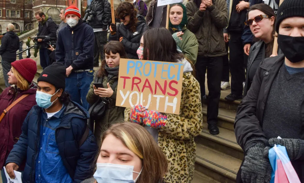 Dans la foule, des militants brandissent des pancartes, l'une d'entre elles disant « protégez les jeunes trans ».