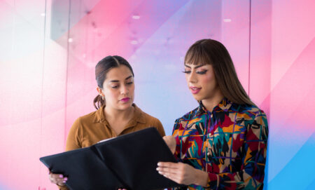 There are two women overlooking a report. One is wearing a brown shirt and the other has a more colourful shirt on. There is a creative background of the trans pride flag colours.