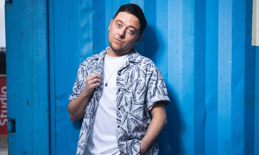 La star d'Emmerdale et acteur trans, Ash Palmisciano, porte une chemise blanche et un bouton à motifs bleus et gris par-dessus alors qu'il regarde vers la caméra.