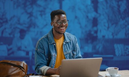 This is an image of a Black man smiling while looking down at his computer. He is wearing a denim shirt with a yellow tshirt underneath.