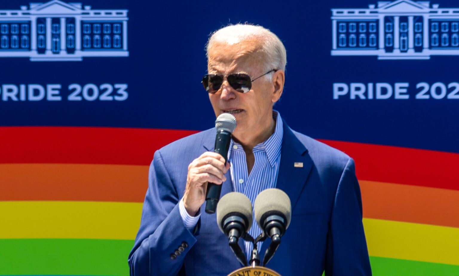 Biden finalise de nouvelles règles radicales et vitales du Titre IX protégeant les étudiants LGBTQ+ de la discrimination President Joe Biden speaking into a microphone, with a picture of a rainbow flag behind him.
