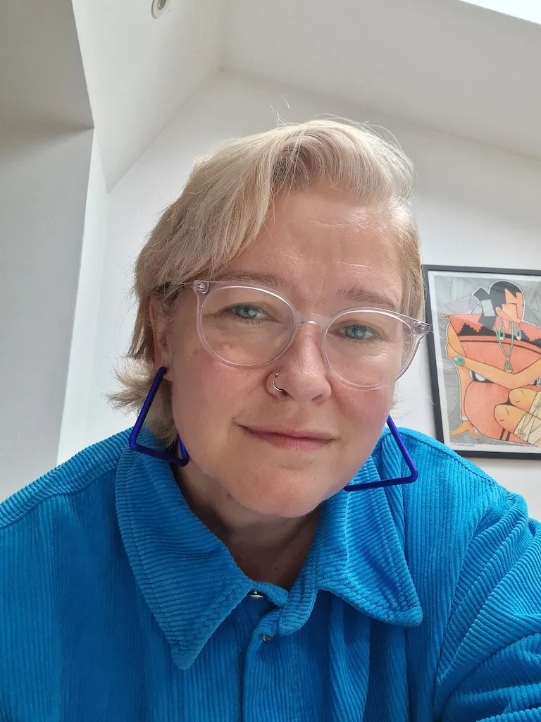 C'est l'image d'une femme.  Elle a les cheveux blonds plus courts et porte des lunettes à monture transparente.  Elle porte une chemise bleue à col montant
