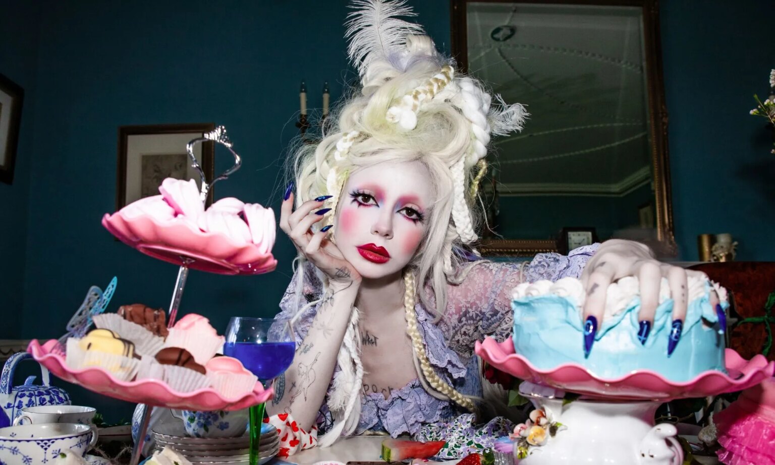 Le message simple de la star non binaire de l'Eurovision, Bambie Thug, pour les haineux : « Faites preuve d'auto-compassion » Bambie Thug in white face makeup and a towering blonde wig resting their hand on their chin.