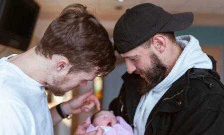 Two caucasian men in their thirties are affectionally holding and looking at a newborn baby lovingly.