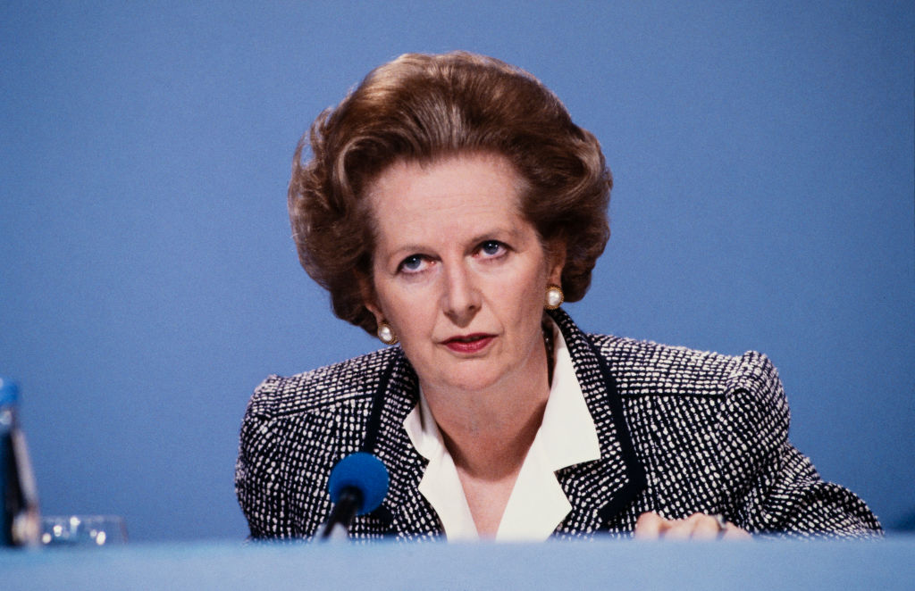 Margaret Thatcher dans un costume gris et une chemise blanche, assise devant un fond bleu