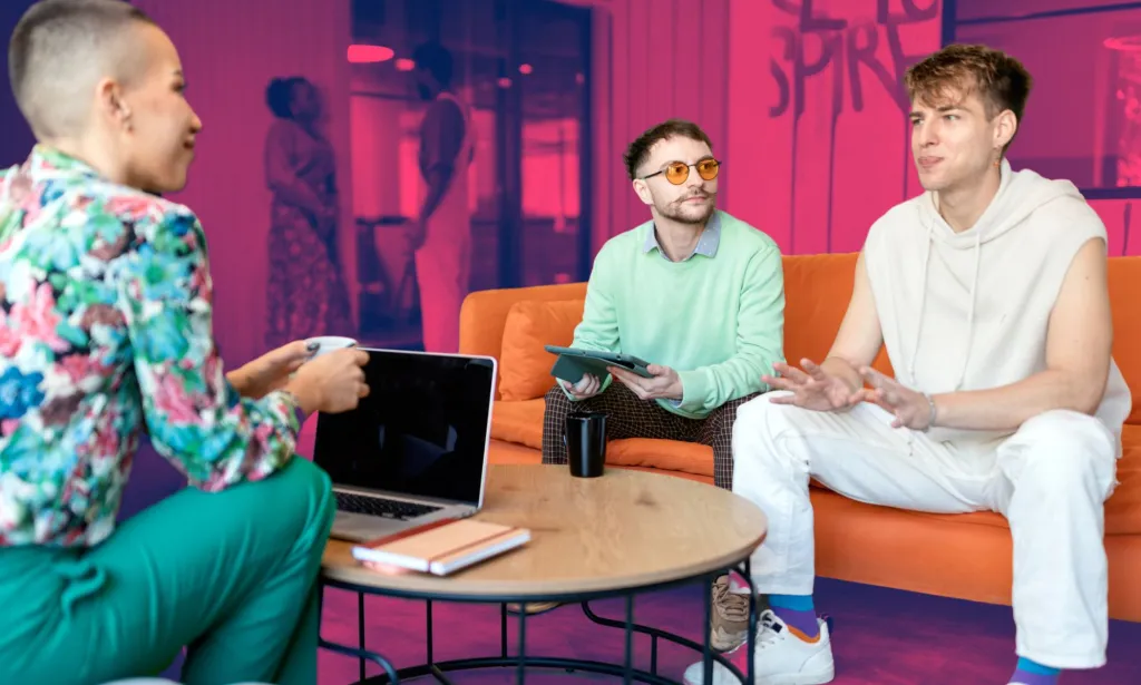 Il s'agit d'une image de 3 employés de Gen réunis dans une zone commune de leur bureau. Ils sont tous habillés de façon vibrante et dialoguent joyeusement les uns avec les autres.