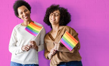This is an image of two queer women holding the Pride flag on a pink wall.
